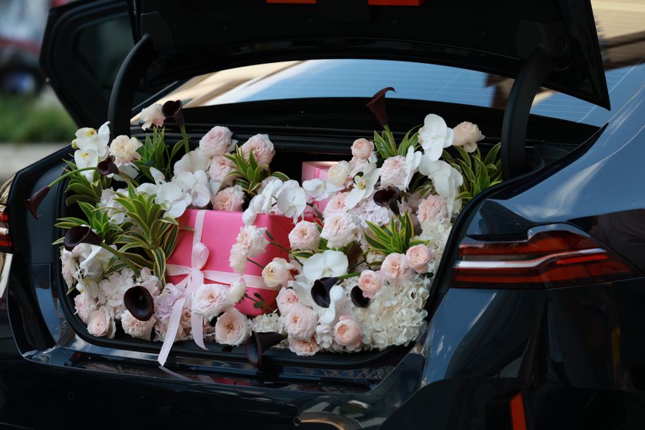 A car's open trunk filled with a vibrant arrangement of fresh flowers, including white orchids, pale pink roses, and dark purple calla lilies, interspersed with green foliage. Among the flowers, there