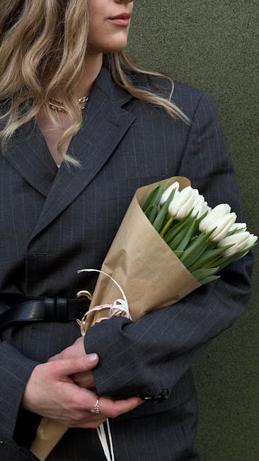 Close-up of a woman wearing a dark pinstripe blazer holding a bouquet of fresh white tulips wrapped in brown craft paper with a white ribbon, standing against a muted green background. The tulips have