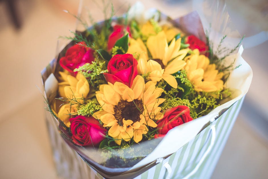 A vibrant floral bouquet featuring bright yellow sunflowers with dark centers, complemented by pink and red roses. The arrangement is wrapped in clear plastic and white tissue paper, with hints of gre