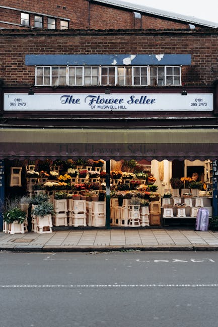 The exterior of 'The Flower Seller' store on Muswell Hill features a large sign with elegant script lettering, situated above a wide display window showcasing a vibrant array of floral arrangements. T