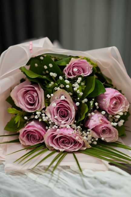 A bouquet of fresh pink roses with tightly curled petals and delicate textures, complemented by small white baby's breath flowers and lush green foliage. The roses are arranged in a compact, rounded s